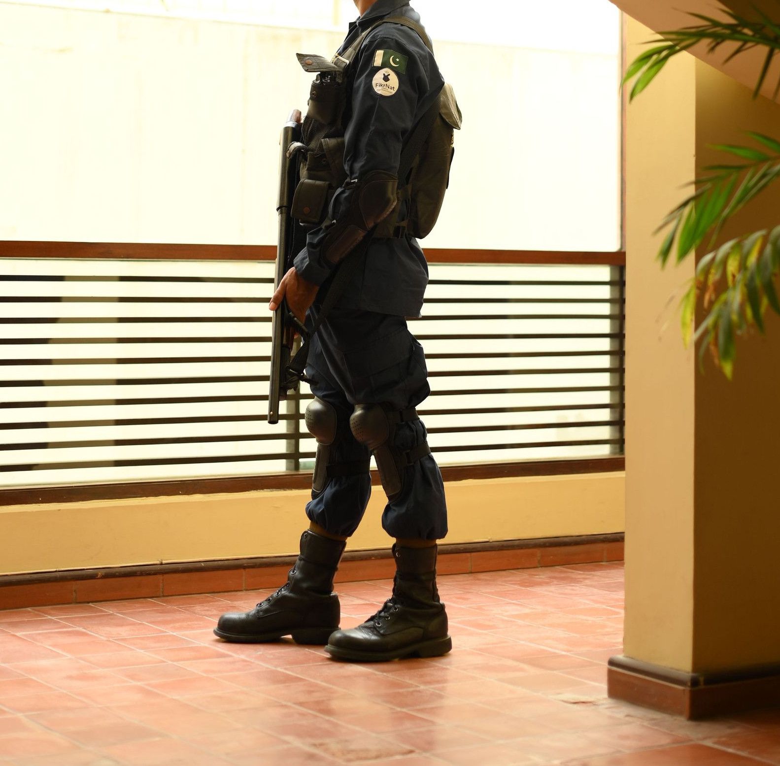 A security guard in uniform stands indoors, observing through a window by a staircase.